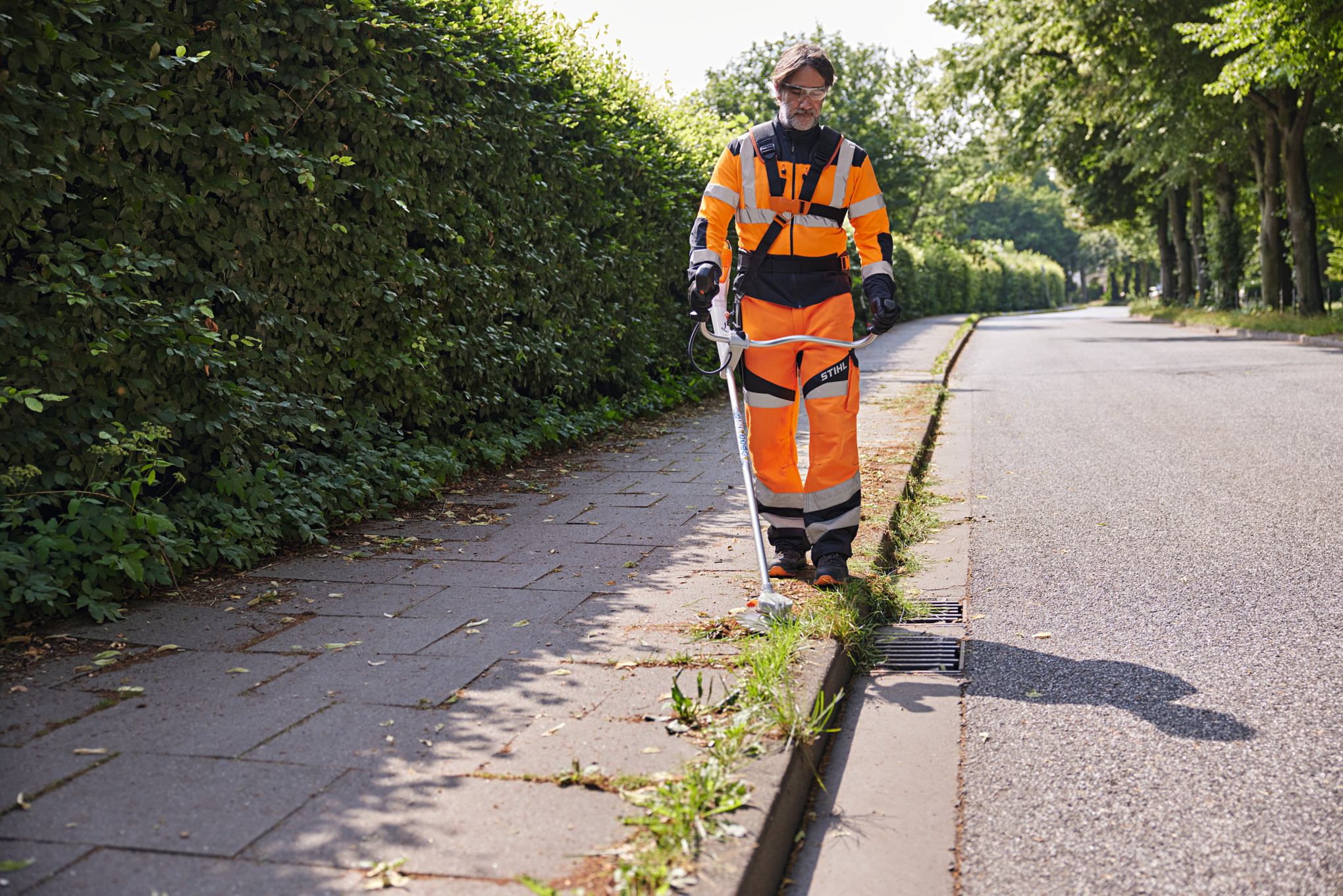 Ein Mann mäht mit dem kabellosen STIHL RGA140 das Gras entlang einer Bordsteinkante in einer Allee