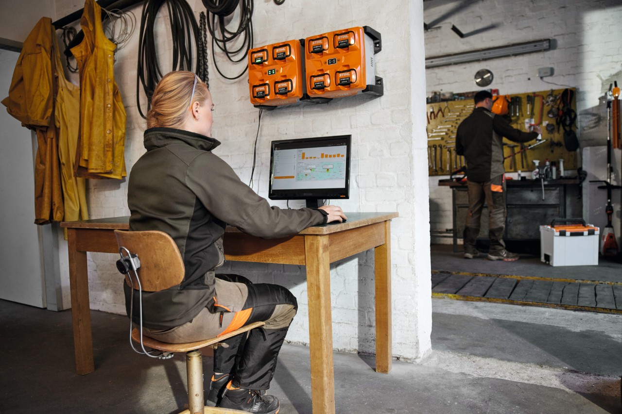 Eine Frau sitzt am PC und arbeitet am Flottenmanagement; über ihr hängt ein Multi-Ladegerät für STIHL-Akkus