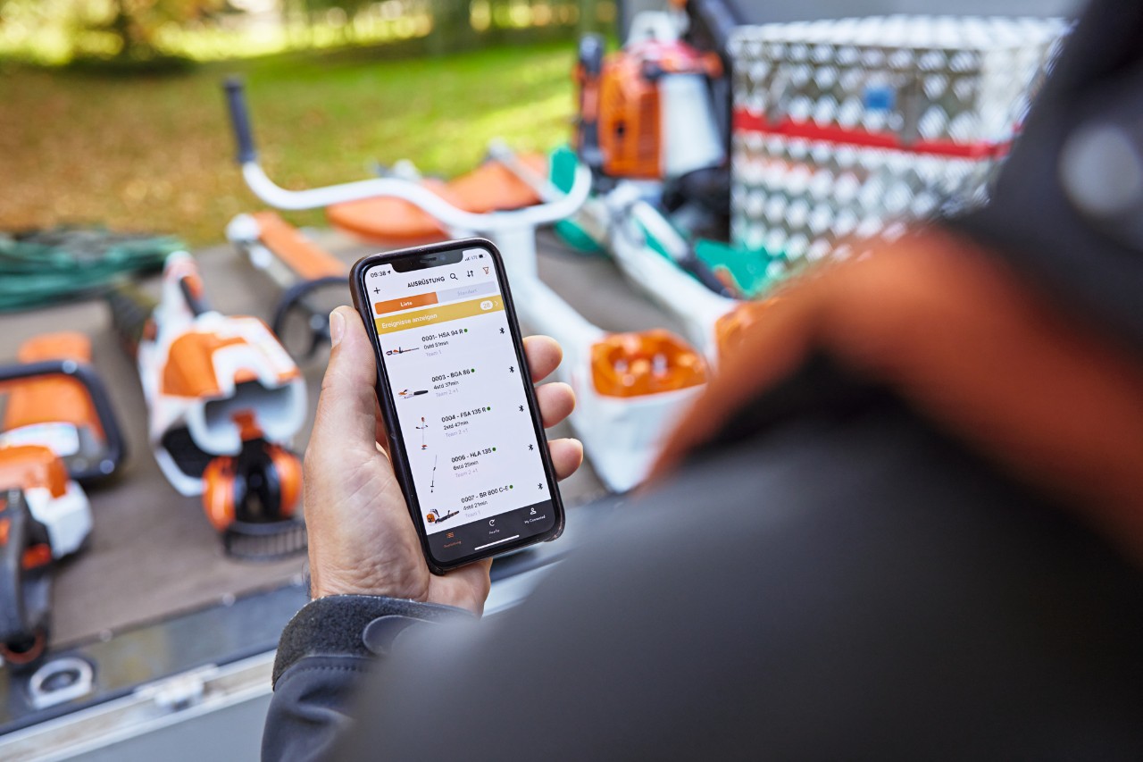 Jemand steht vor der Ladefläche eines Pritschenwagens, auf der diverse STIHL Geräte platziert sind. Die Person hält ein Smartphone in der Hand, auf dem die STIHL connected App geöffnet ist. 