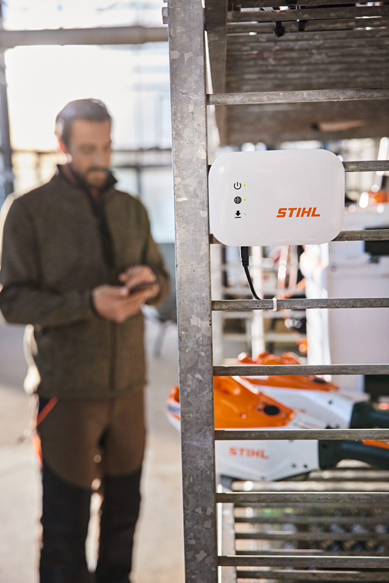 Ein STIHL connected Box hängt an einem Werkstatt-Regal. Im Hintergrund ist ein Garten-und Landschaftsbauer zu sehen, der mit seinem Smartphone interagiert. 