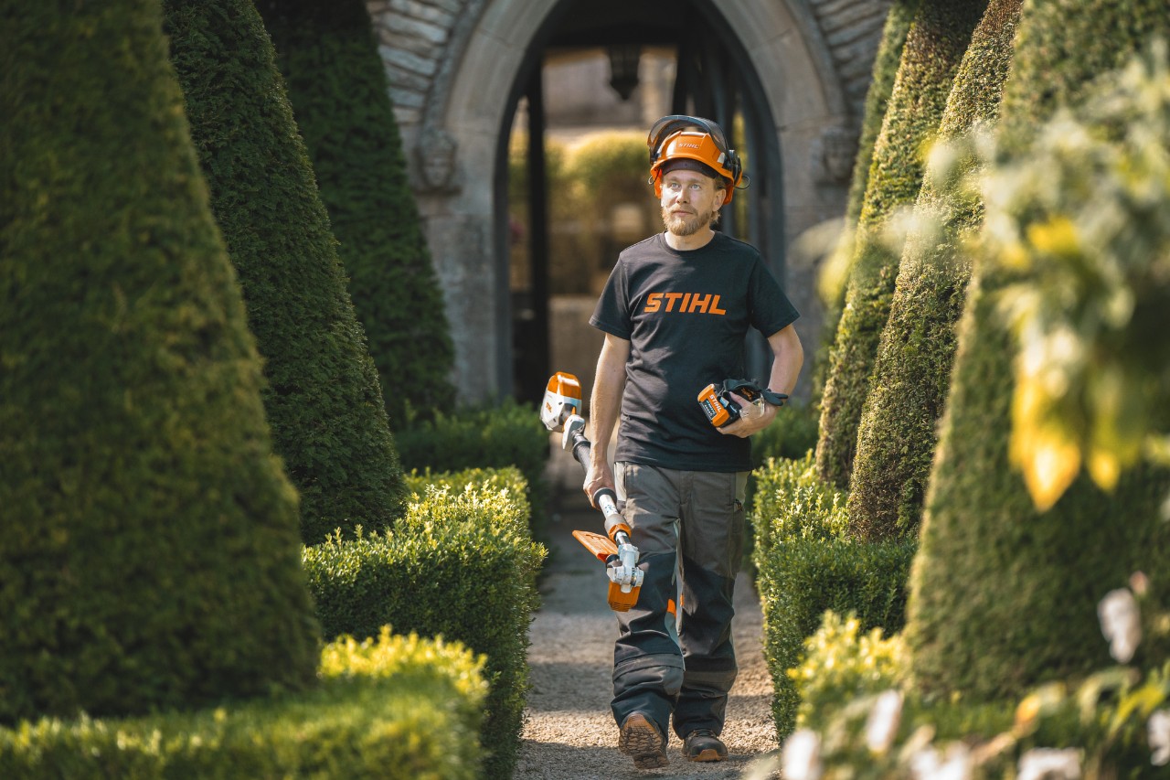 Chefgärtner Andrew Wain läuft mit einem Schutzhelm, einem STIHL T-Shirt und einem STIHL Akku-Gerät mit separatem Akku in der Hand durch einen Gartengang
