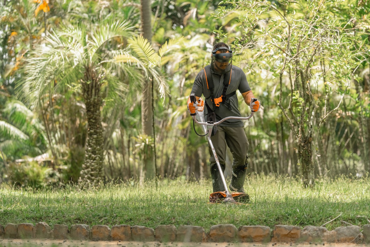 Mitarbeiter des Botanischen Gartens mit Arbeitshandschuhen und Schutzhelm bei der Arbeit auf dem Rasen mit einer Motorsense 