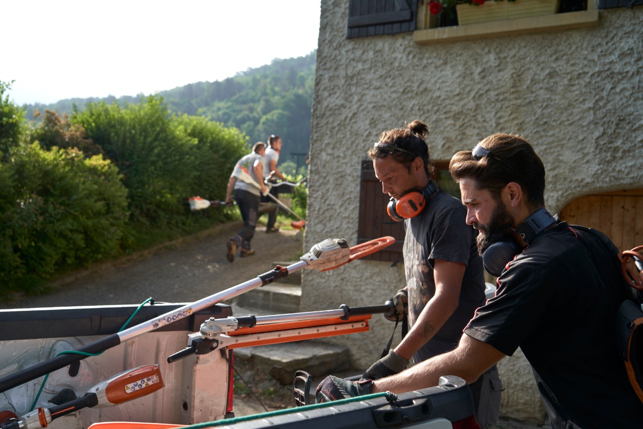 Zwei Landschaftsgärtner entladen STIHL Akku-Geräte aus einem Auto, im Hintergrund sind weitere Arbeiter 