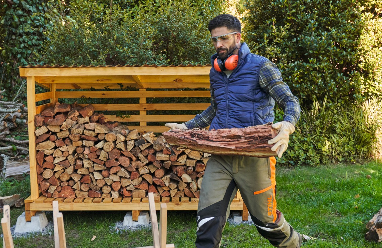 Ein Mann mit Schutzbrille trägt Feuerholz, im Hintergrund ein Brennholzunterstand