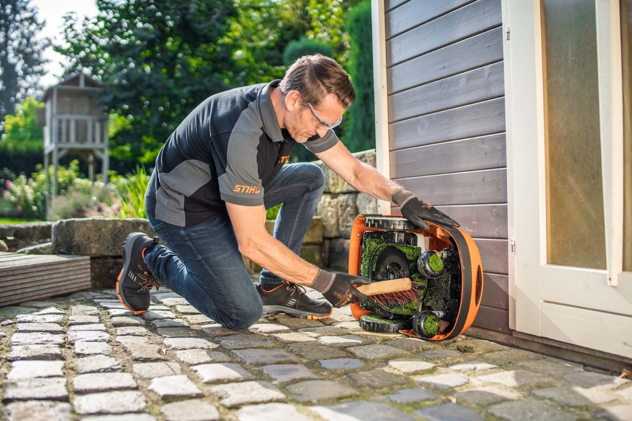 Mann reinigt den STIHL iMOW® Mähroboter 422  von unten mit einer Bürste, um ihn danach winterfest zu machen