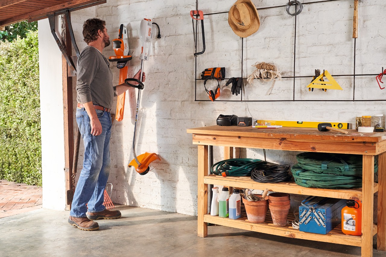 Ein Mann in einer Garage greift nach einer STIHL Elektro-Motorsense an der Wand, daneben eine STIHL Akku-Heckenschere