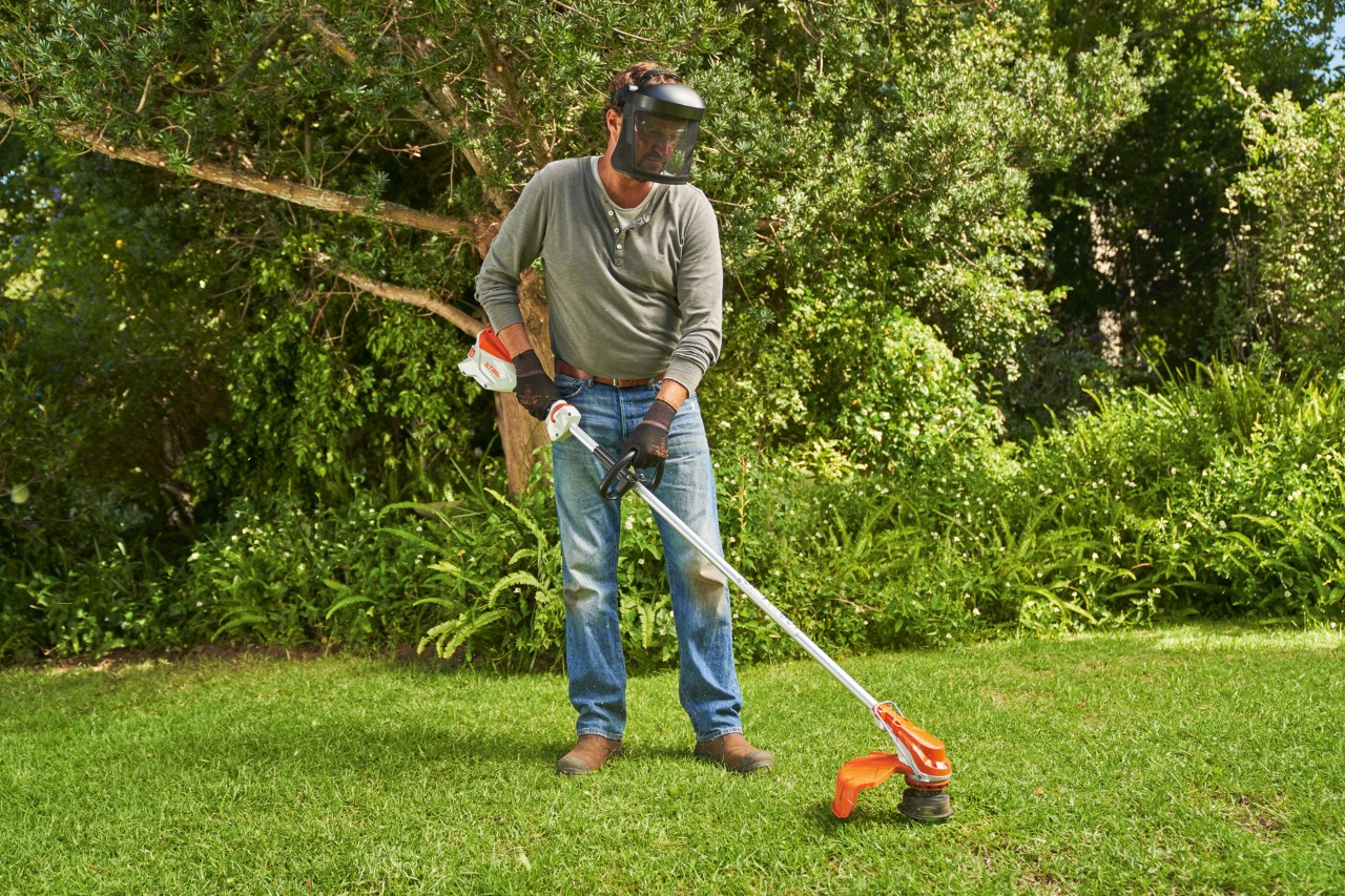 Ein Mann mit Gesichtsschutz ist dabei, in einem Garten mit der Akku-Motorsense STIHL FSA 60 den Rasen zu mähen