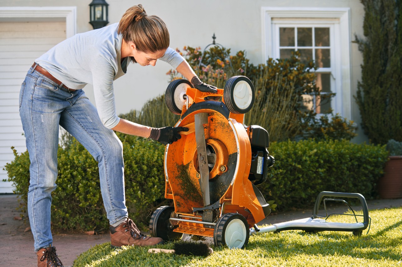 Frau reinigt die Unterseite eines STIHL Benzin-Rasenmähers RM 448 T-C mit einem Spatel