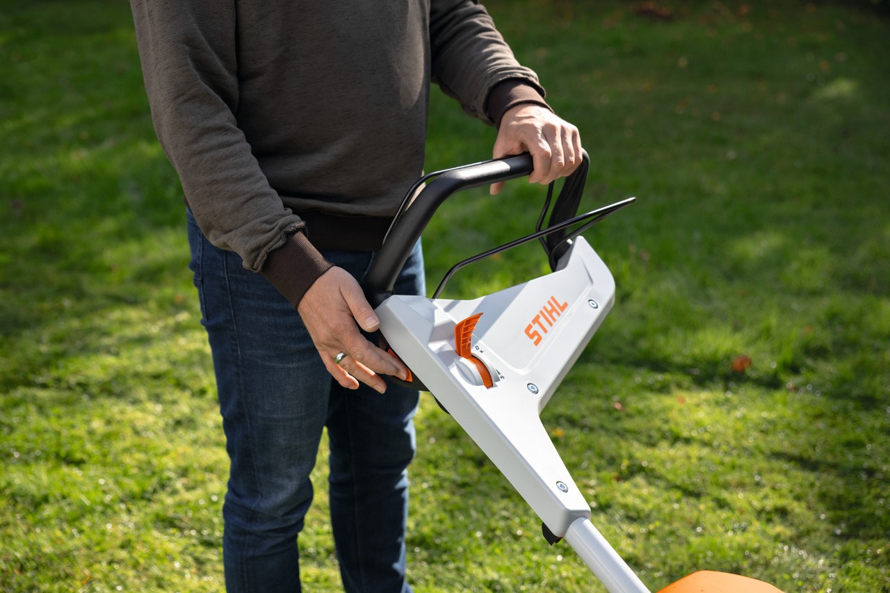 Eine Person auf einer Wiese drückt den Starterknopf eines Akku-Rasenmähers STIHL