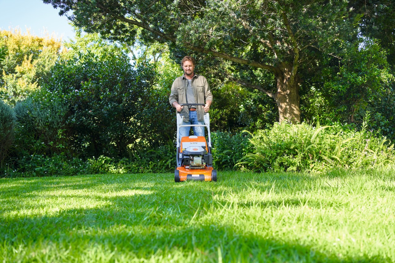 Ein Mann mäht mit einem STIHL Rasenmäher einen halbschattigen Rasen, im Hintergrund Büsche und Bäume