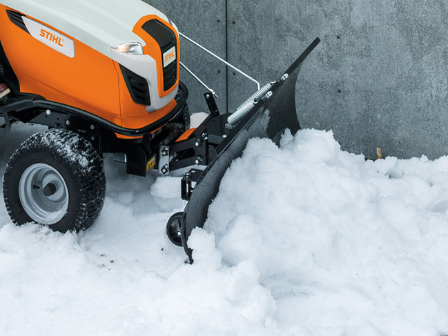 Mit einem Schneeschild an einem Rasentraktor wird Schnee vor einer Mauer geräumt