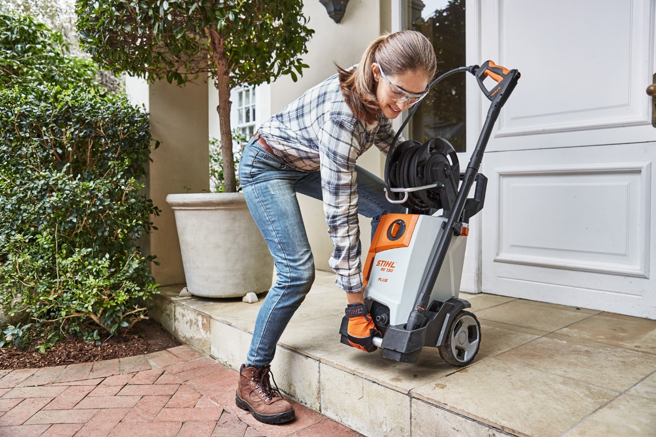 Frau hievt einen STIHL Hochdruckreiniger RE 130 plus die Stufe vor einer Wohnungstür hinunter