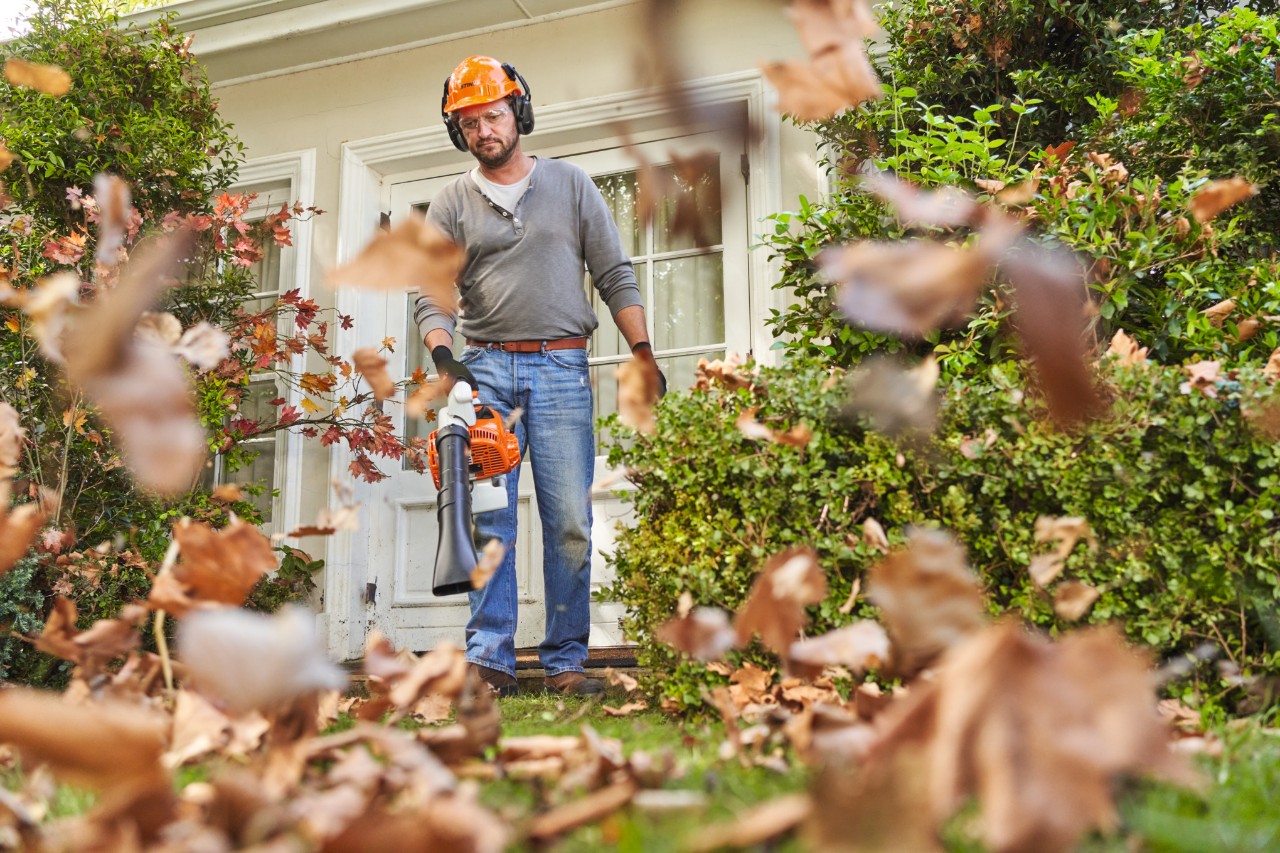Ein Mann in STIHL Schutzausrüstung vor einem Haus pustet Blätter mit dem Blasgerät weg