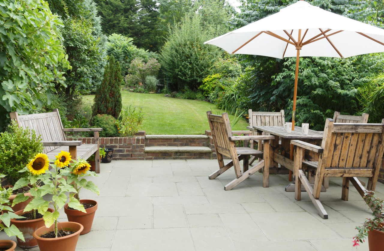 Holzmöbel auf einer geräumigen Terrasse mit Betonsteinen an einem großen, grünen Garten