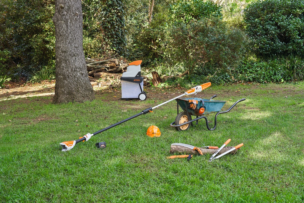 Verschiedene Geräte, zum Beispiel eine STIHL Garten- und Astschere, liegen auf einer Rasenfläche in einem Garten, um einen Apfelbaum zu schneiden