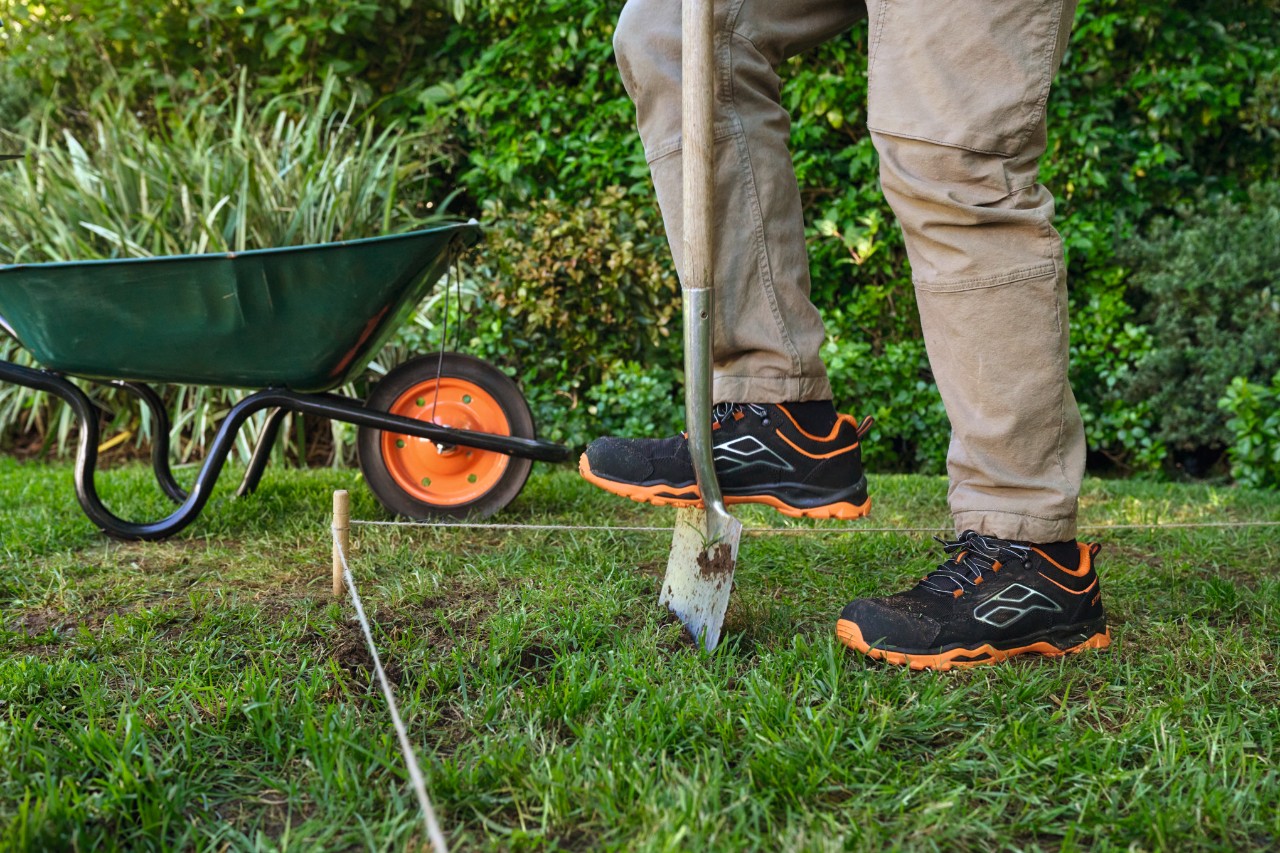Nahaufnahme, wie ein Mann mit Arbeitshandschuhen die Grasnarbe mit einem Spaten etwa 5 Zentimeter tief aushebt