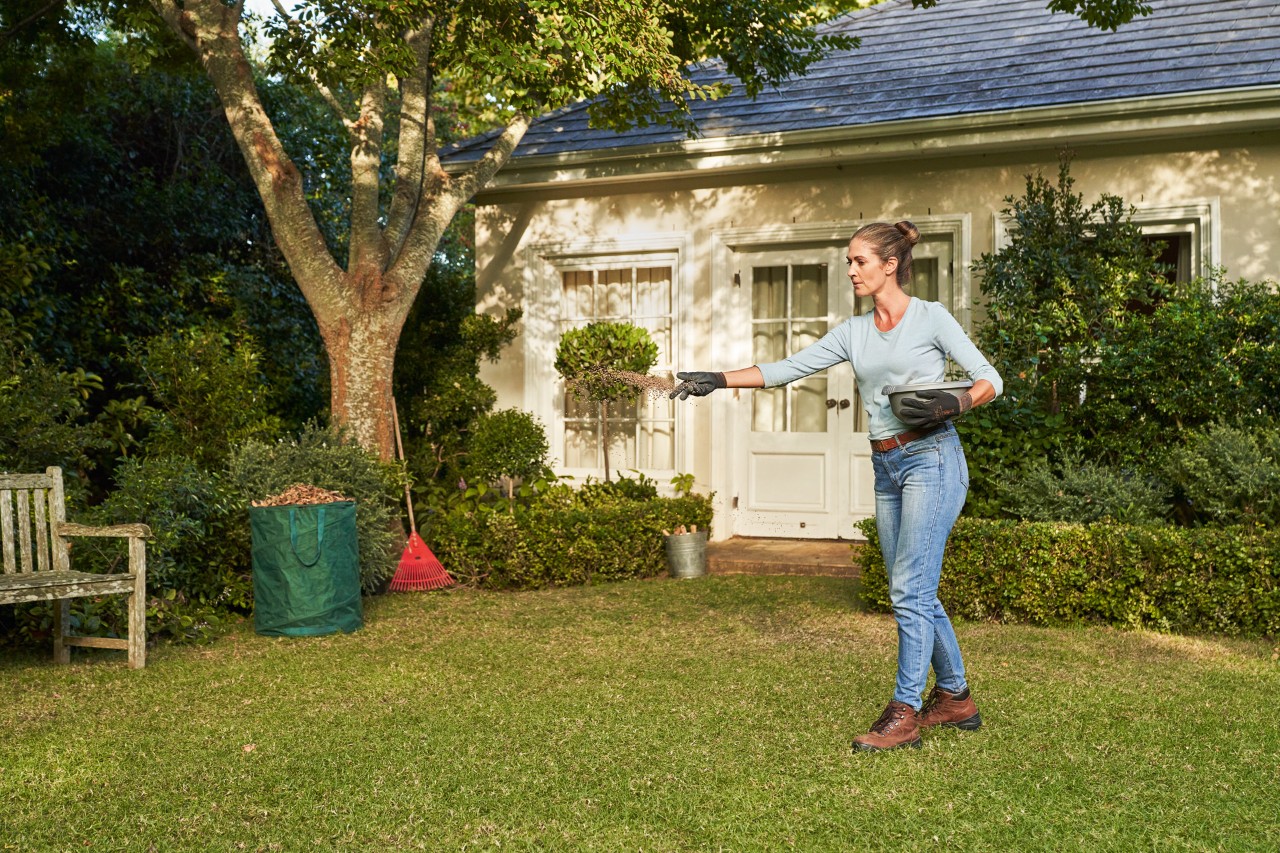 Eine Frau mit Handschuhen STIHL FUNCTION SENSOTOUCH bringt Gartenkalk auf dem Rasen aus, im Hintergrund Büsche und ein Haus