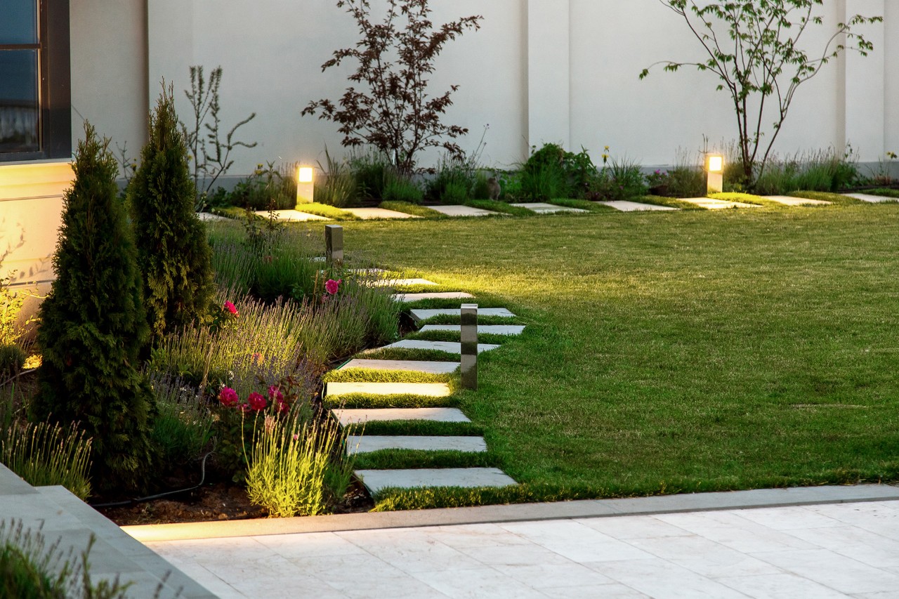 Kleiner Garten abends, Blumenbeet, grüner Rasen, Marmorsteg aus quadratischen Fliesen, dekorative Bodenlampen-Beleuchtung.