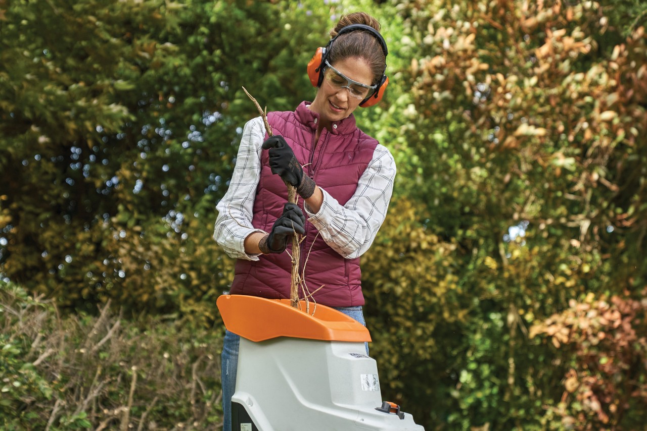 Eine Frau in Schutzausrüstung entsorgt Schnittgut aus dem verwilderten Garten mithilfe eines Elektro-Gartenhäckslers STIHL GHE 140