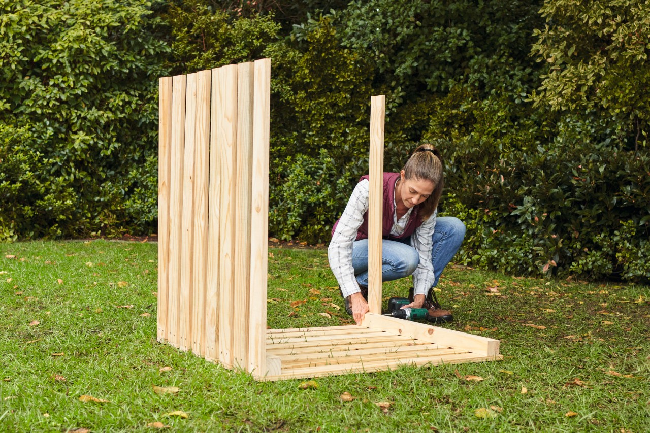 Frau mit Jeans und Pferdeschwanz befestigt mit einem Akkuschrauber auf einem Gartengrundstück die Latten für die Seitenwände am Komposter