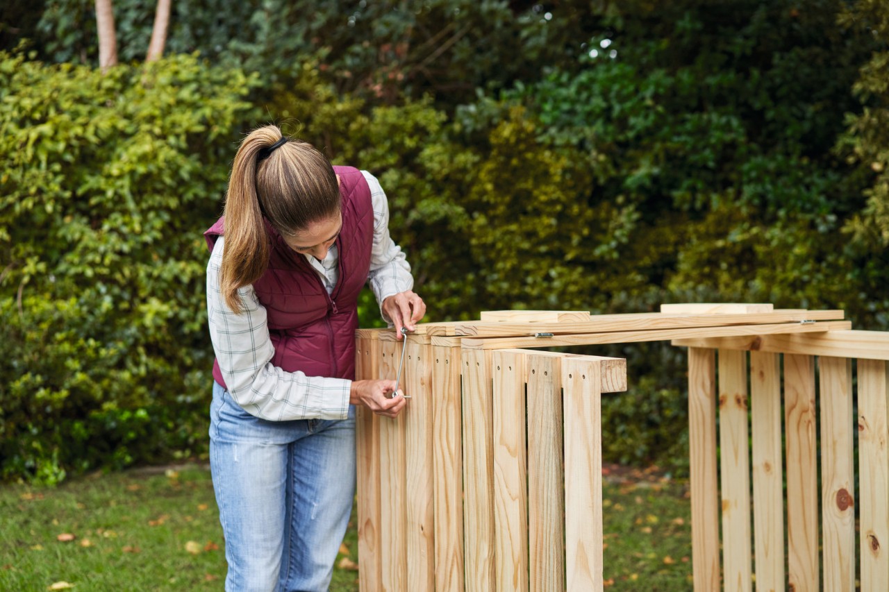 Frau auf einem Gartengrundstück befestigt Sturmhaken am selber gebauten Komposter 