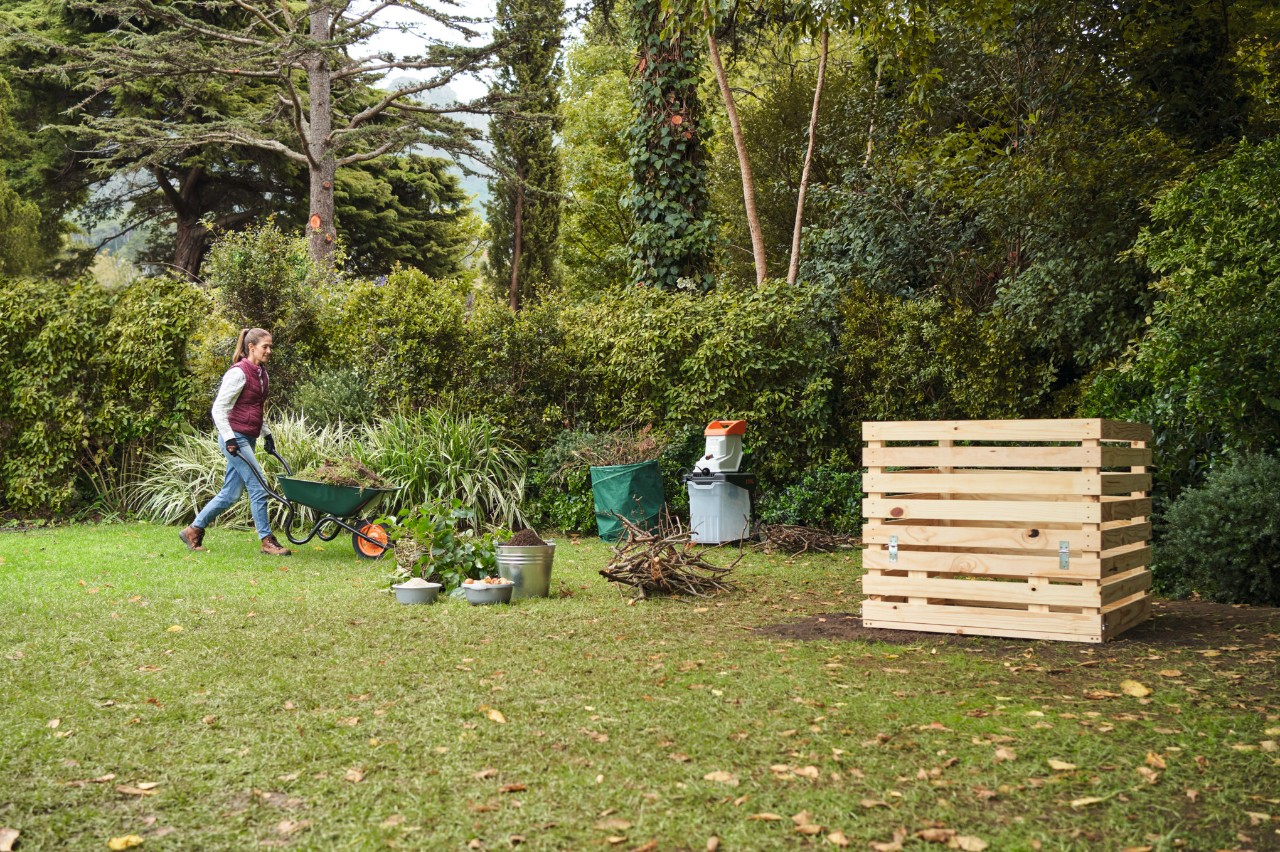 Eine Frau auf einem Gartengrundstück steuert mit einer Schubkarre auf einen selbstgebauten Holzkomposter zu