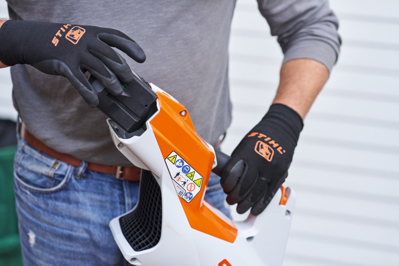 Person mit schwarzen STIHL Handschuhen entnimmt einem Blasgerät den Akku