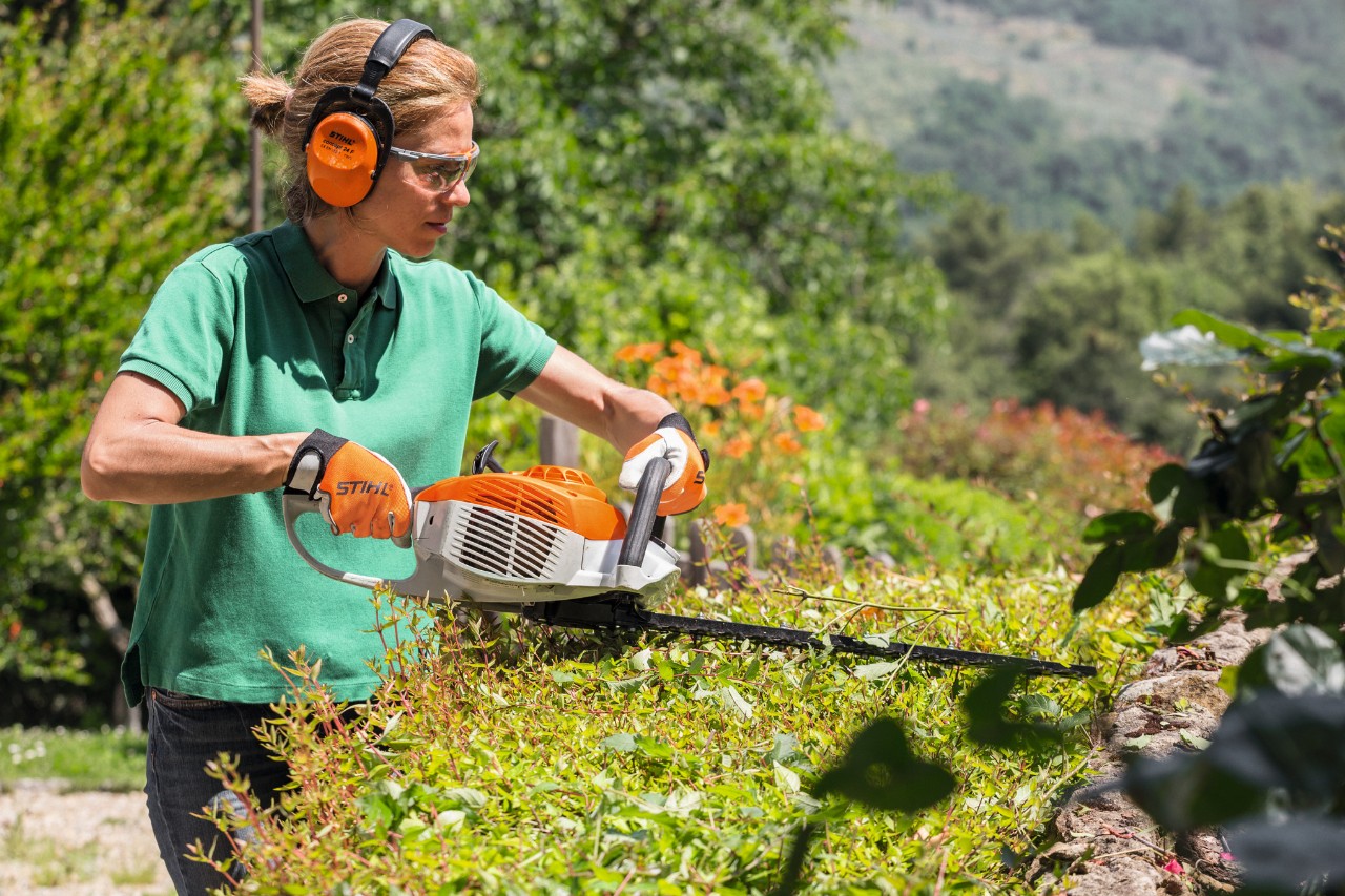 Frau mit Gehörschutz und Schutzbrille schneidet mit einer STIHL Benzin-Heckenschere HS 46 eine Hecke