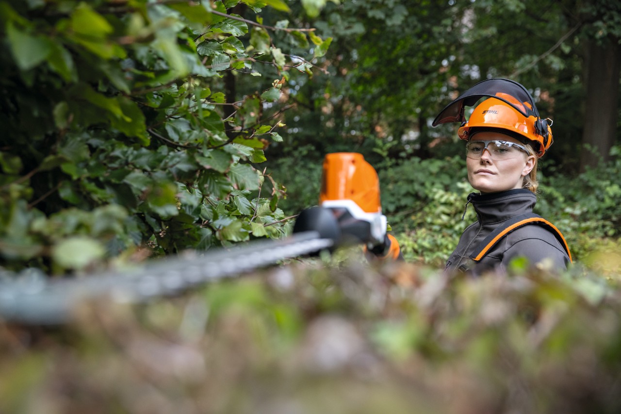 Blonde Frau in Schutzausrüstung beim Schneiden einer Buchenhecke