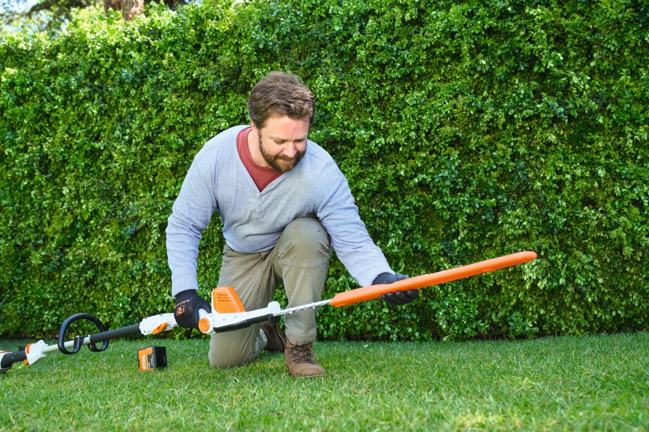 Ein Mann vor einer Hecke legt einen Akku-Heckenschneider STIHL HLA 56 auf dem Rasen ab