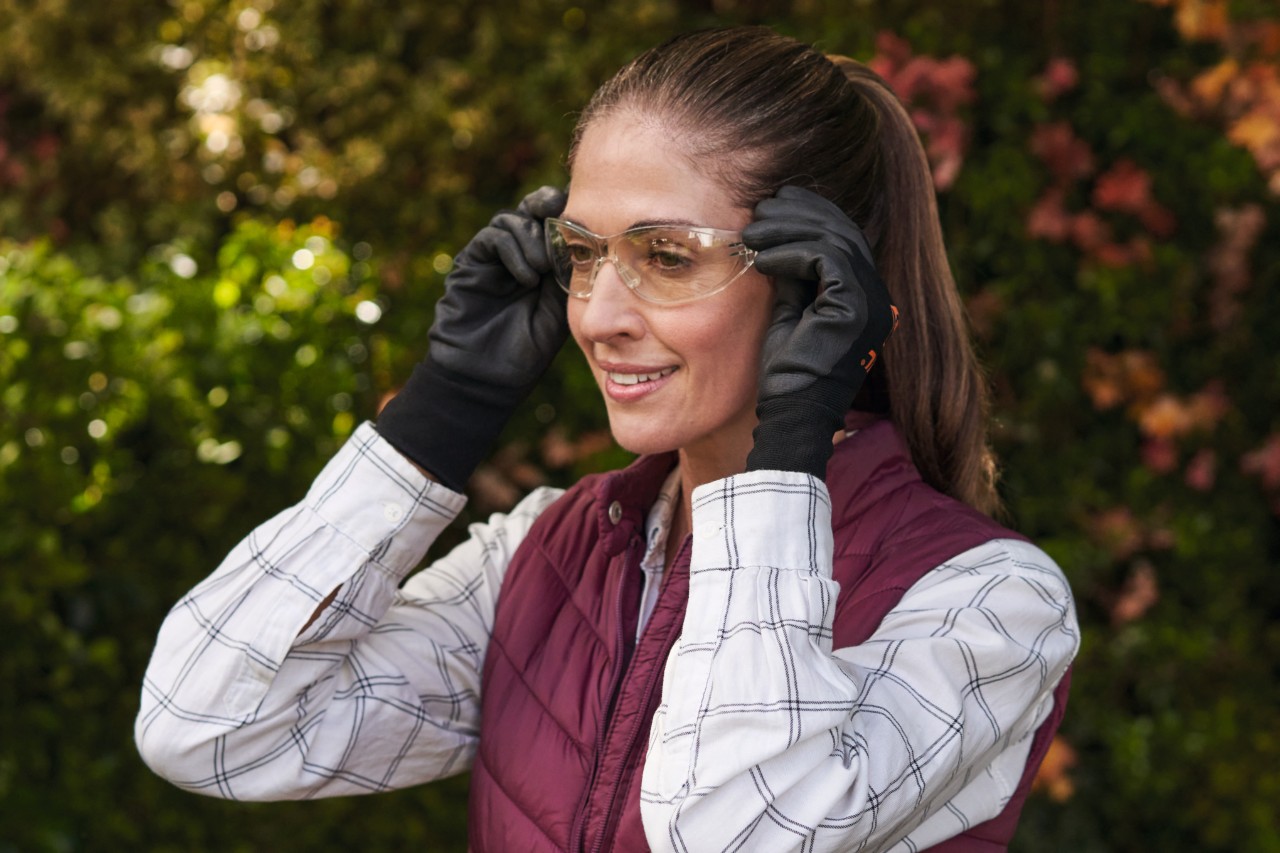 Lächelnde, braunhaarige Frau mit Schutzhandschuhen setzt sich eine Schutzbrille auf 