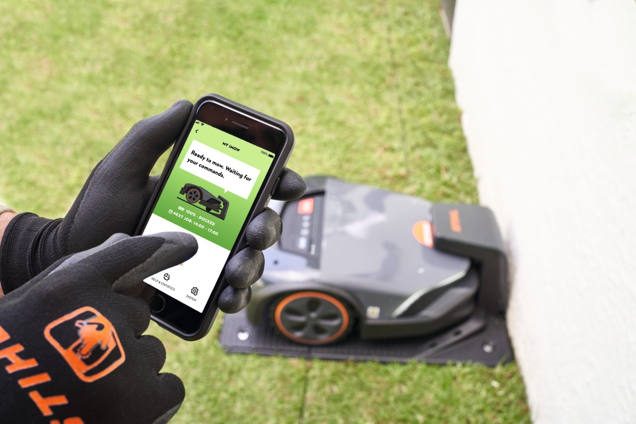 Jemand hält ein Smartphone mit geöffneter My iMOW App in der Hand, auf dem Display erscheint die Abbildung eines Rasenmähers und in einer Sprechblase der Text: “Ready to mow. Waiting for your commands.”