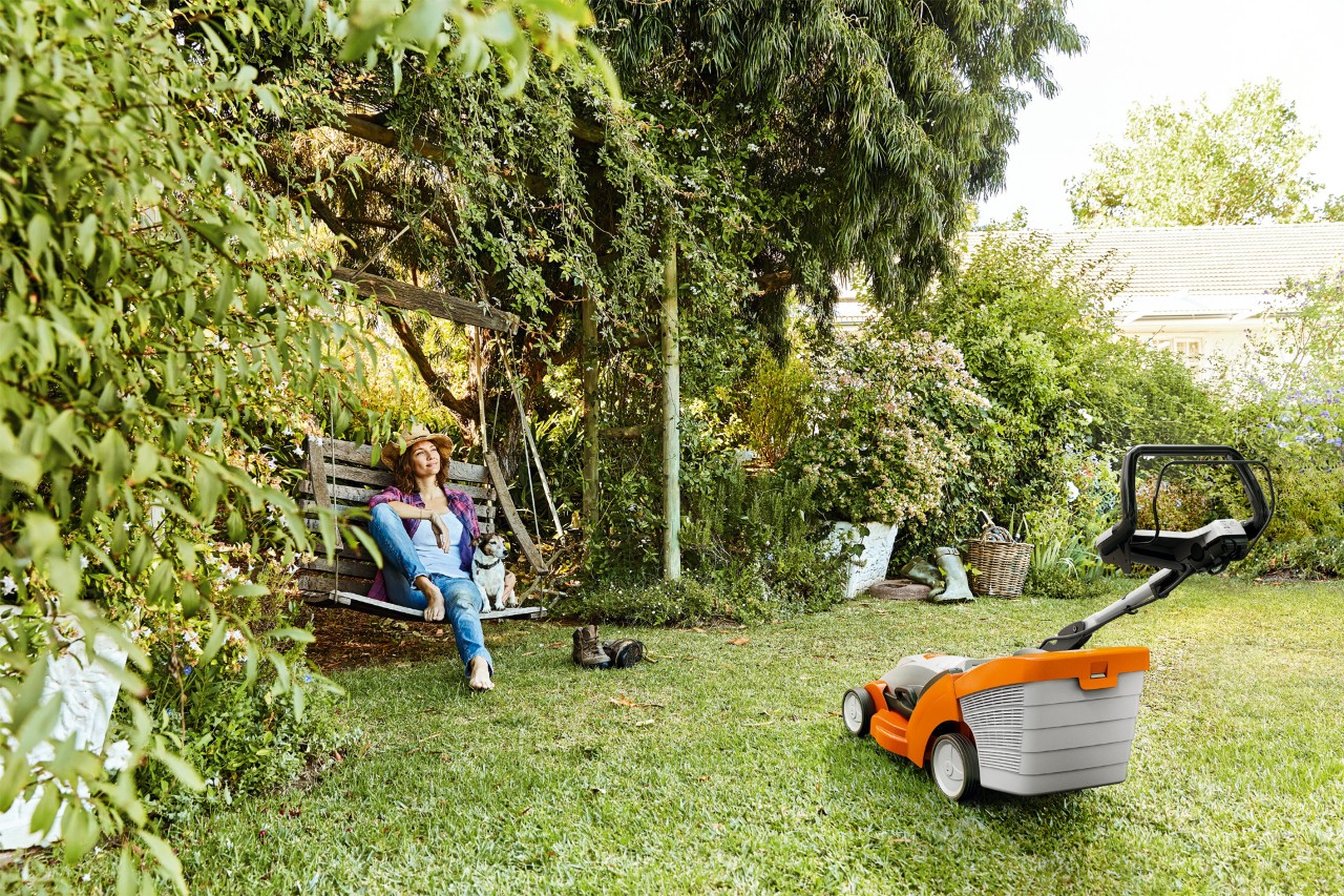 Frau entspannt auf einer Gartenschaukel neben einem STIHL Akku-Rasenmäher RMA 339 auf einem grünen Grundstück
