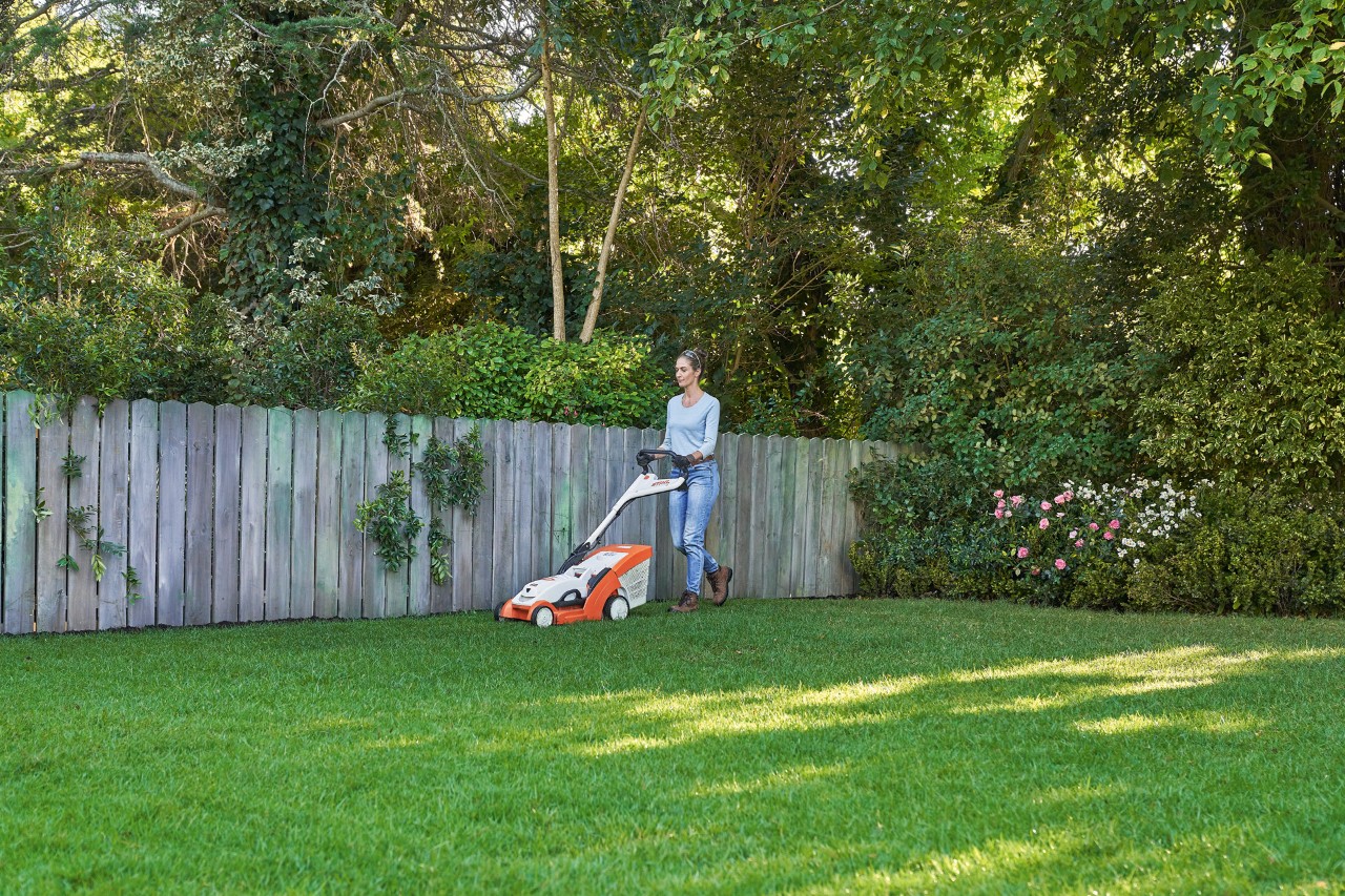 Frau mäht grüne Rasenfläche vor einem Holzzaun mit STIHL Akku-Rasenmäher RMA 339 C.