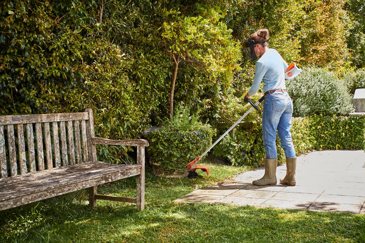 Frau trimmt in einem Garten neben einer Bank mit dem STIHL Akku-Freischneider FSA 57 Rasenkanten an Büschen