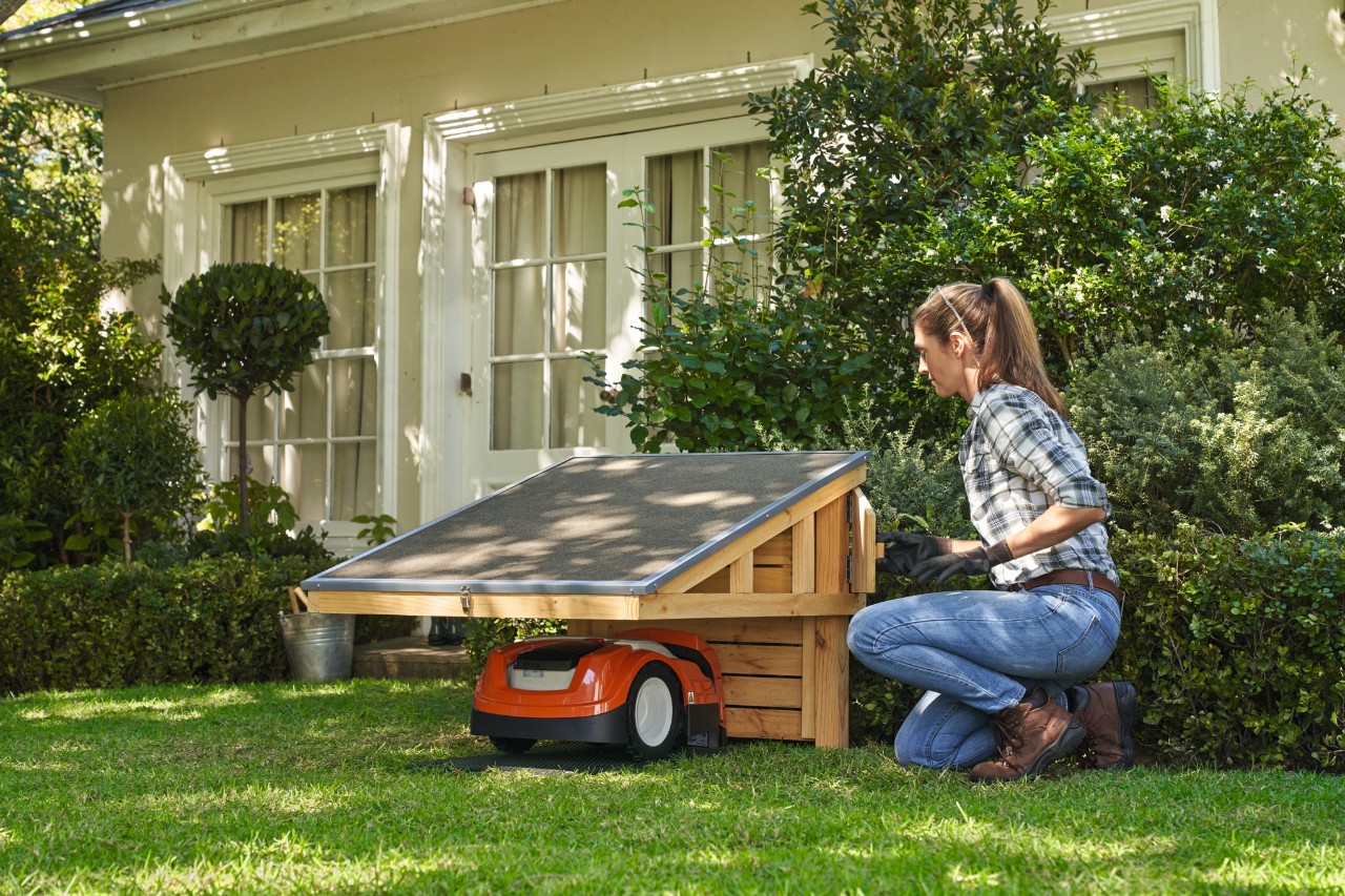 Frau mit Schutzbrille und Arbeitshandschuhen kniet auf Rasen neben  STIHL iMOW®-Garage mit geparktem STIHL iMOW® Mähroboter, öffnet Ablagefach