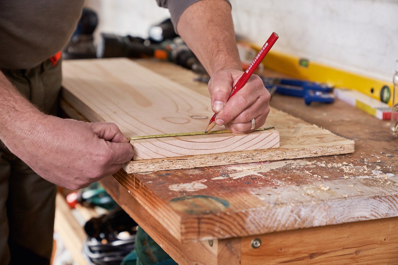 Ein Mann markiert Bohrlöcher auf einem Holzbrett, um einen Gartengerätehalter selber zu bauen