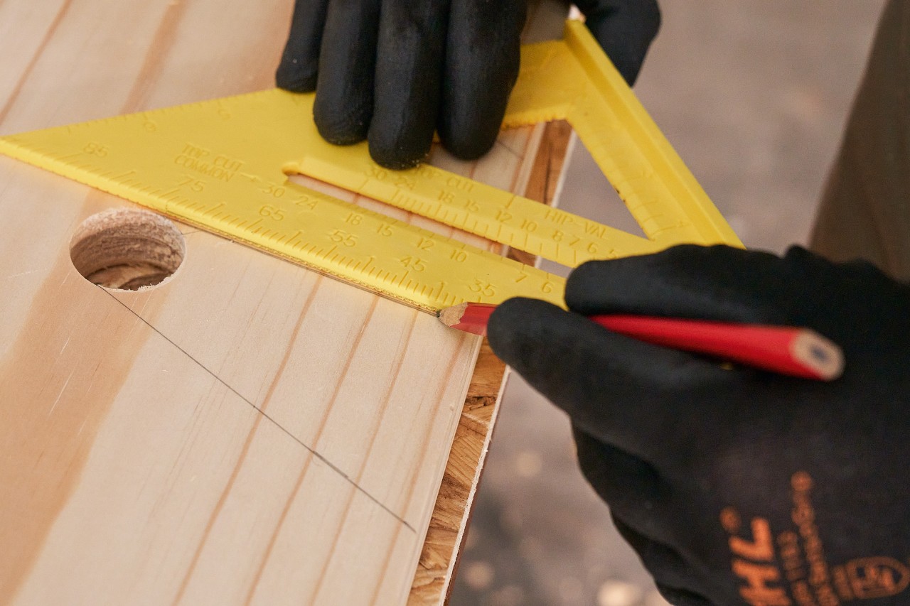 Jemand zeichnet mit Bleistift und Geodreieck Linien auf ein Holzbrett, um Aussparungen zu markieren 