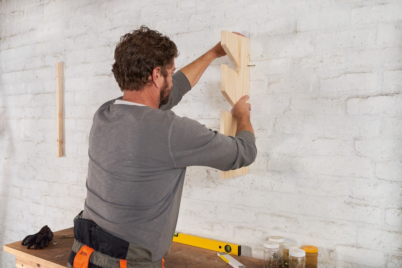 Ein Mann dübelt eine Holzleiste an die Wand, um einen Gartengerätehalter selber zu bauen 
