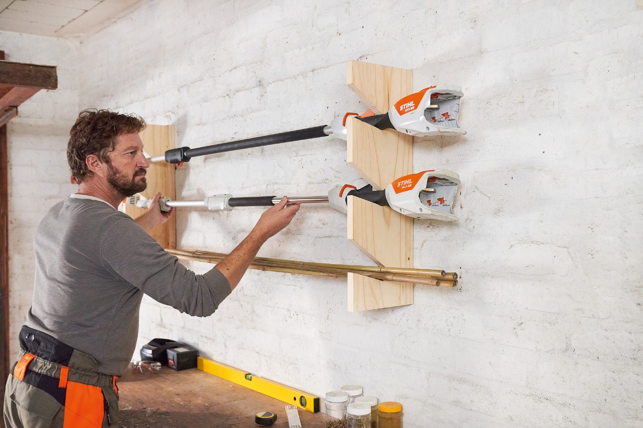 Ein Mann legt eine STIHL Motorsense in einen selber gebauten Gartengerätehalter, befestigt an der Wand 