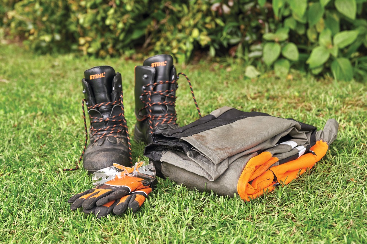 Stiefel, Schutzhandschuhe und Schnittschutzhose liegen auf dem Rasen