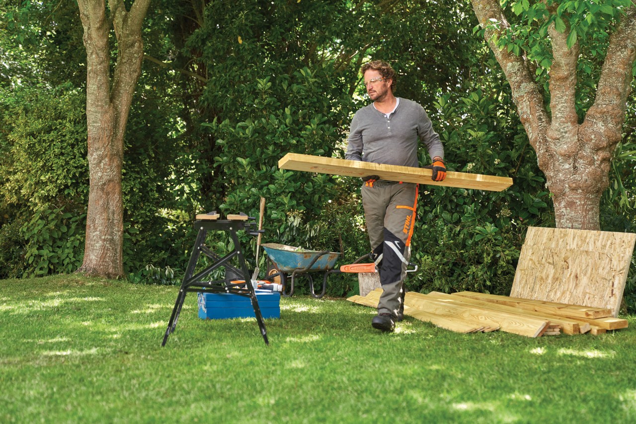 Person nimmt sich ein Holzbrett von einem Stapel unter einem Baum, um einen Geräteschuppen selber zu bauen