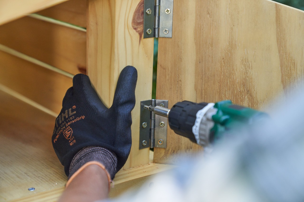 Nahaufnahme: Hand mit STIHL Arbeitshandschuh FUNCTION SENSOTOUCH schraubt Tür vom Staufach der Mähroboter-Garage fest