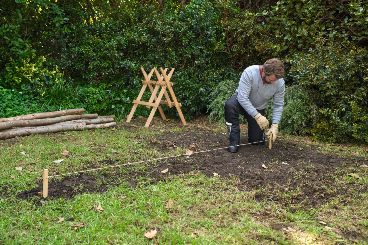 Ein Mann steckt Hölzchen, die mit einer Pflanzschnur verbunden sind, zur Flächenbegrenzung in den Boden