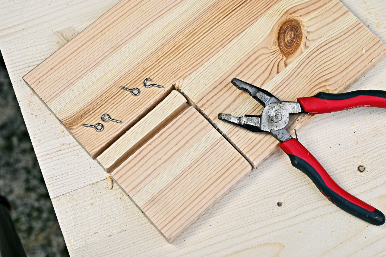 Vier Ösen und eine Zange auf einem Holzbrett, um Hängetür für ein Igelhaus zu bauen