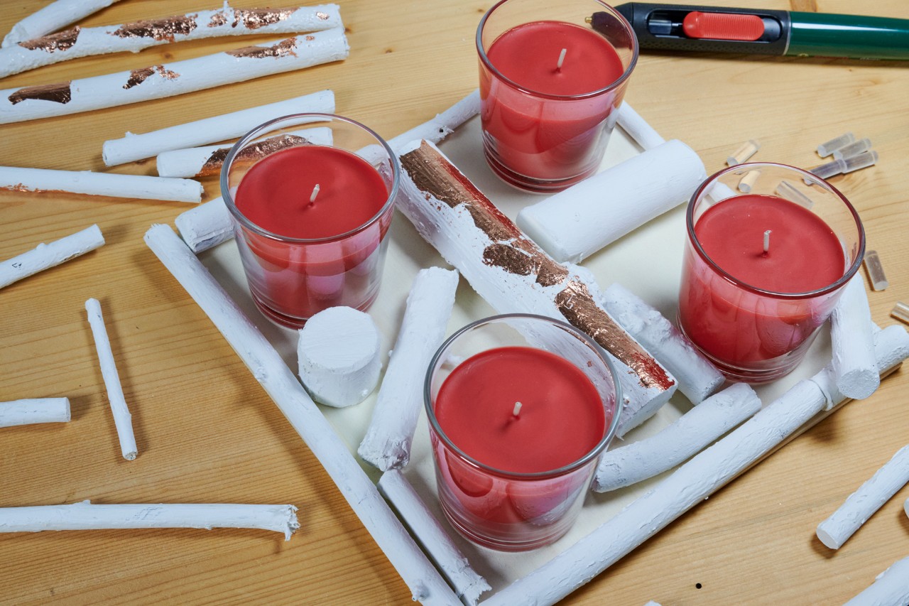 Weiße Äste liegen zwischen vier roten Teelichtern auf einer quadratischen, weißen Holzplatte 