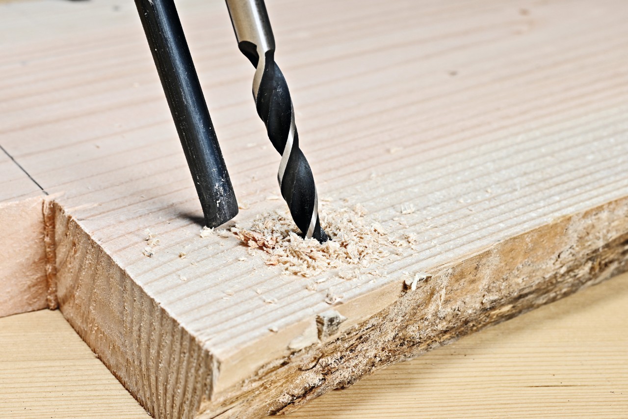 Mit einem Holzbohrer wird ein Loch in die Holzbohle gebohrt, daneben die passende Metallstange