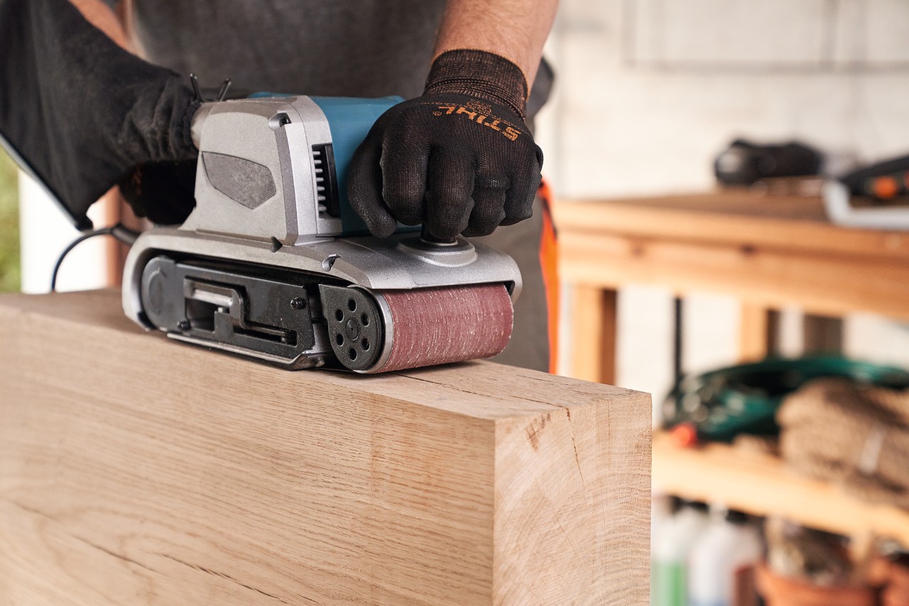 Nahaufnahme einer Person mit STIHL Schutzhandschuhen beim Holzschleifen mit Schleifmaschine in der Werkstatt