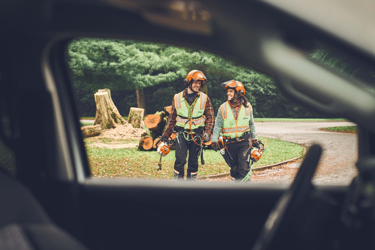 Ein Mann und eine Frau mit STIHL Akku-Motorsägen und einem STIHL Akku-Gehölzschneider laufen lächelnd in Richtung eines Autos  