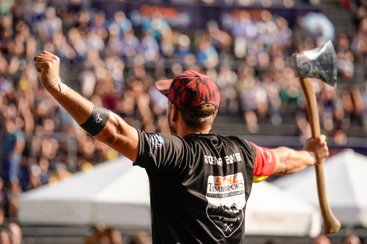 Mann mit STIHL TIMBERSPORTS® Trikot, triumphierend geballter Faust und Axt in der rechten Hand vor Wettkampfpublikum